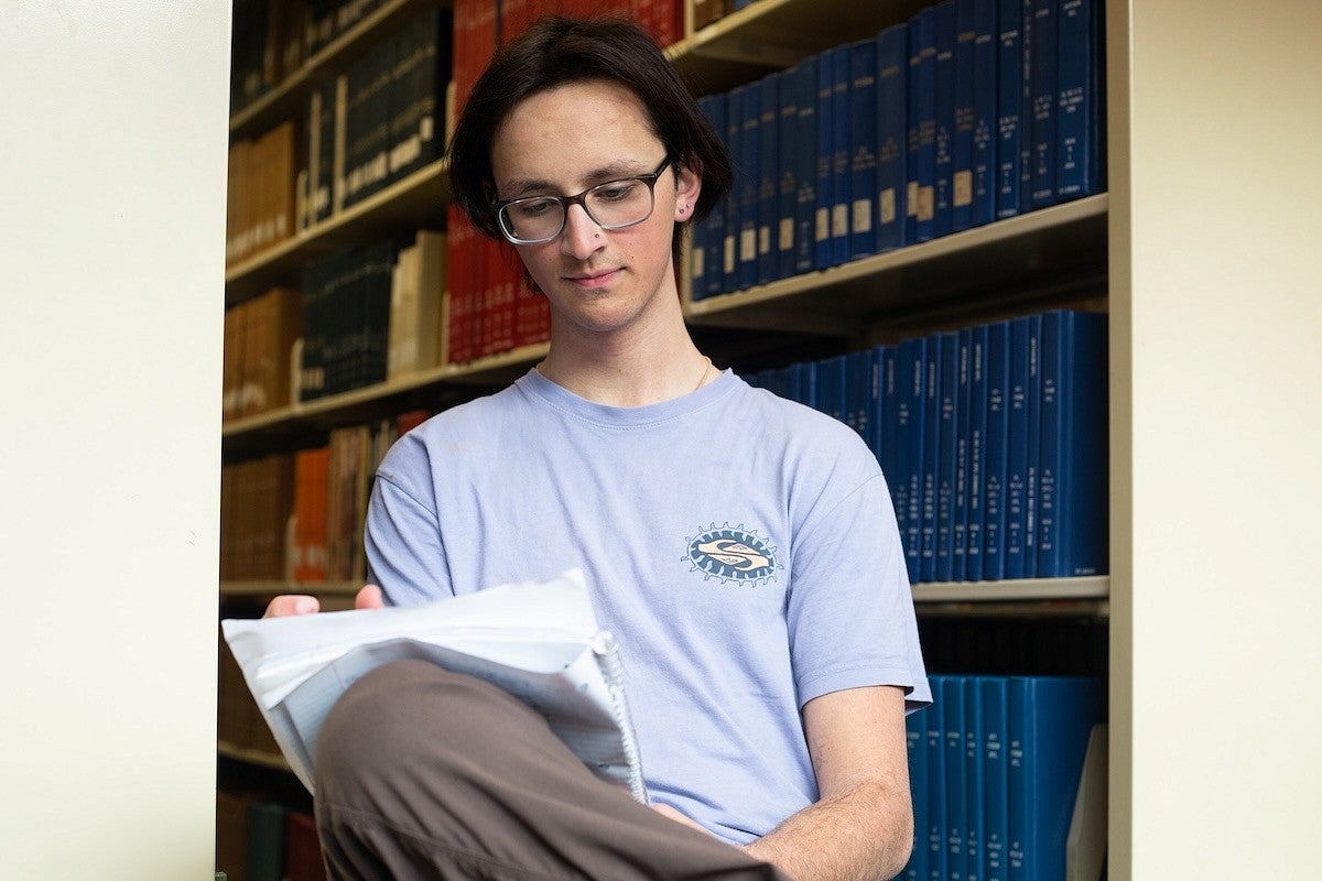 student writing in notebook seated among library stacks