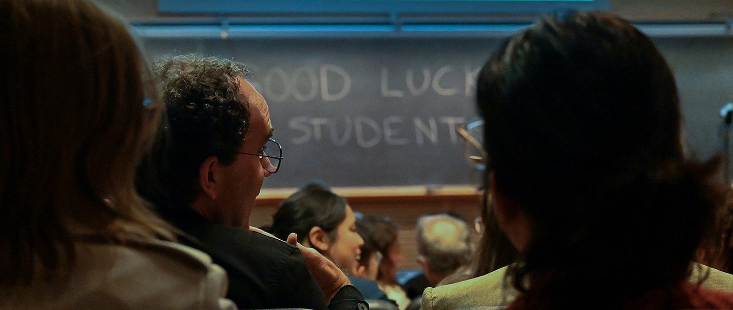 audience members look toward the front of a lecture hall with the words "GOOD LUCK STUDENTS" written on a chalkboard