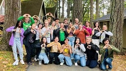 group of students posing among cabins in the woods and throwing the O