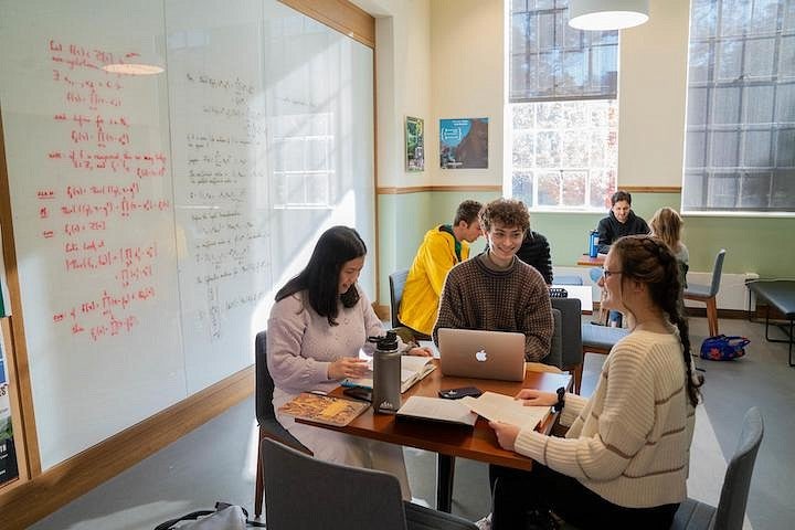 students working around table in light-filled room in front of whiteboard covered in equations