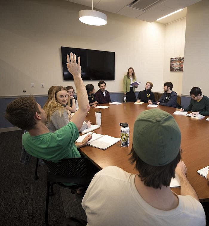 A student in a group of students seated around a class table raises a hand.