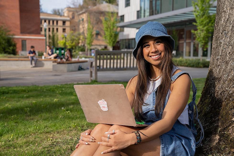 Student with a laptop on her knees sitting against a campus tree.
