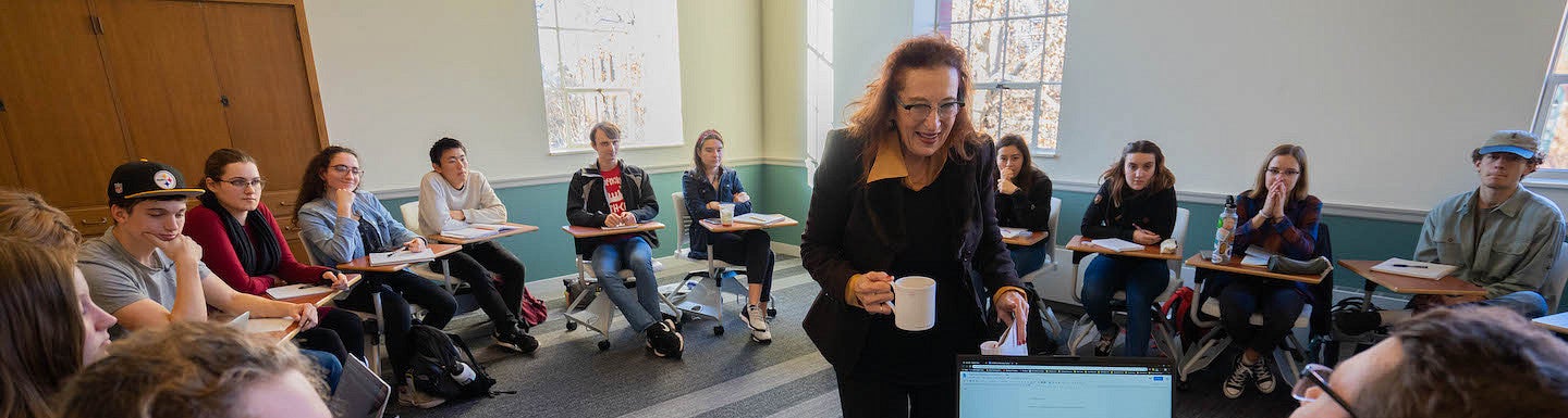 a female faculty member smiling in the center of a ring of students sitting at desks in a light-filled classroom