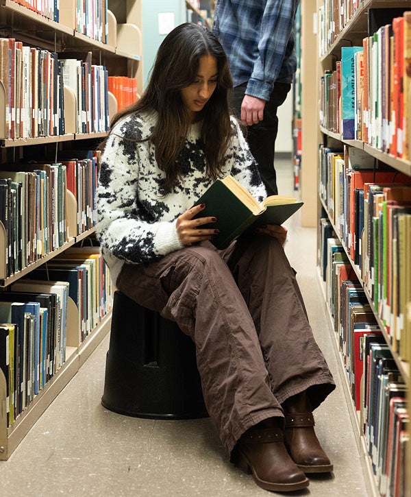 Student sitting on a stool in the library stacks and reading a book