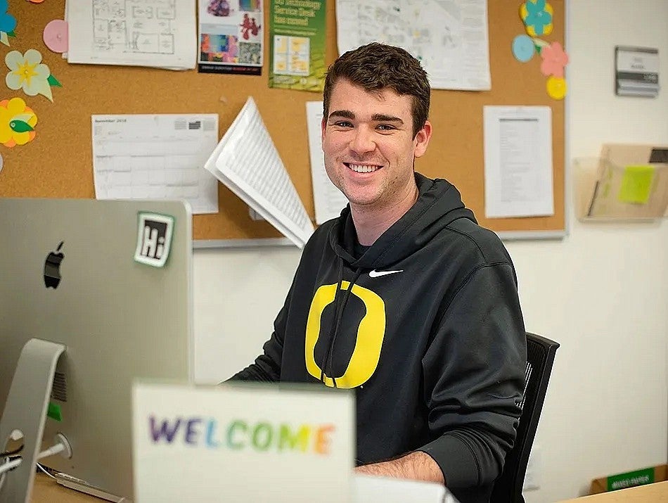 smiling man in a University of Oregon sweatshirt behind a 'welcome' sign