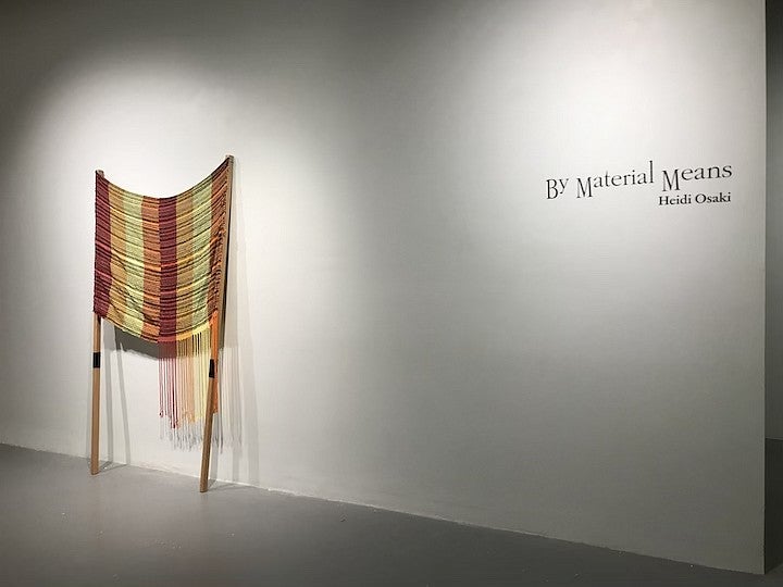 A woven fiber object displayed against a white exbition space wall near text reading "by material means, Heidi Osaki"