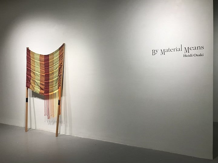 graduate thesis show A woven fiber object displayed against a white exbition space wall near text reading "by material means, Heidi Osaki"