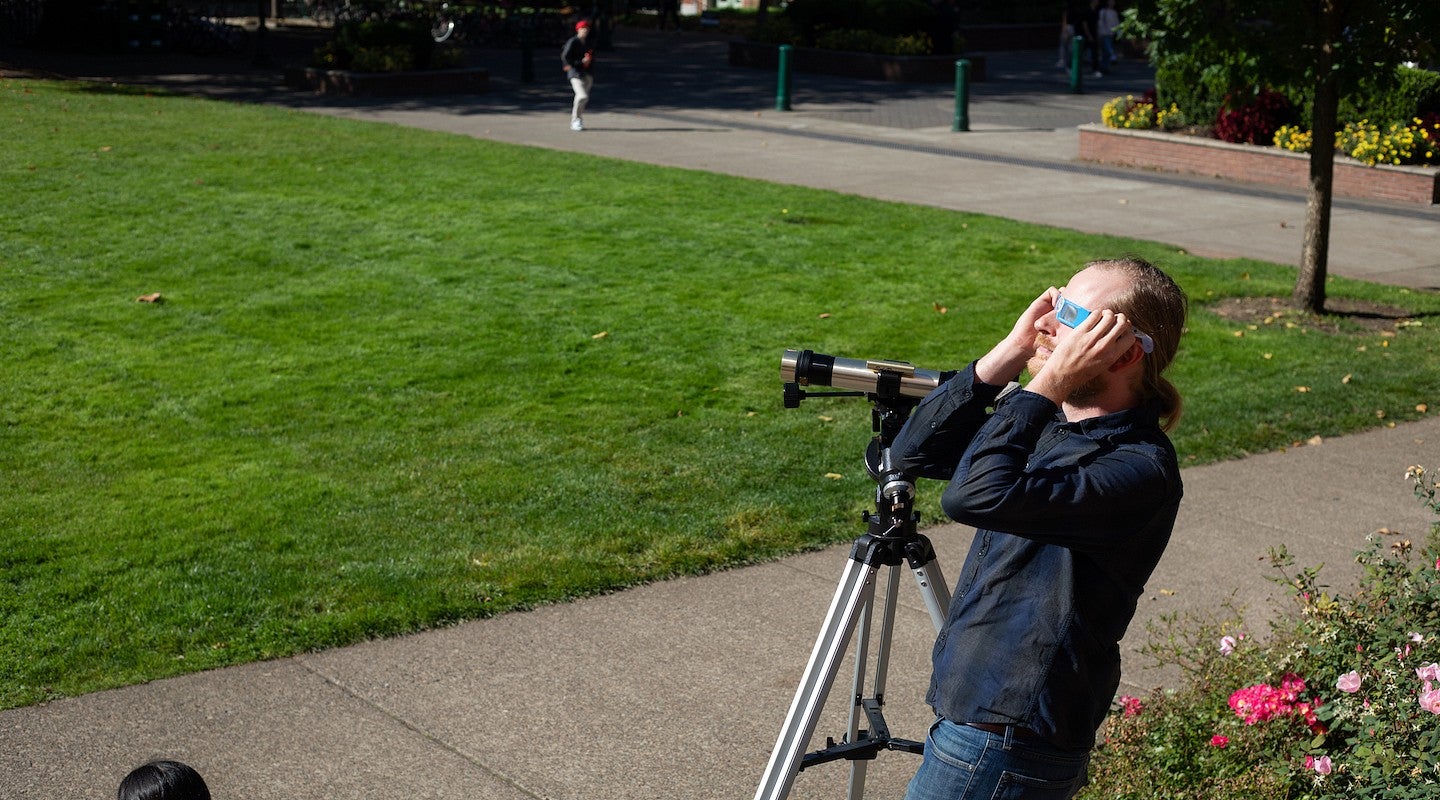 jesse feddersen with telescope on UO green, looking at the sky with eclipse glasses on