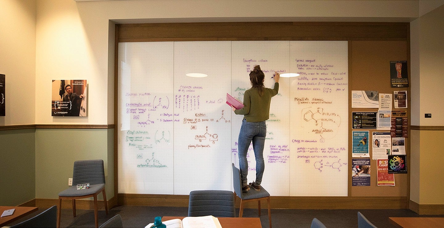 student standing on chair to write equations on large whiteboard wall in Chapman Hall