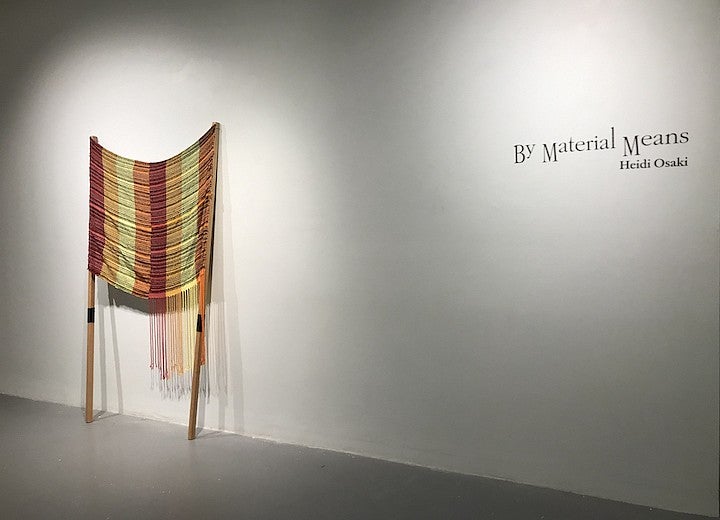 A woven fiber object displayed against a white exbition space wall near text reading "by material means, Heidi Osaki"
