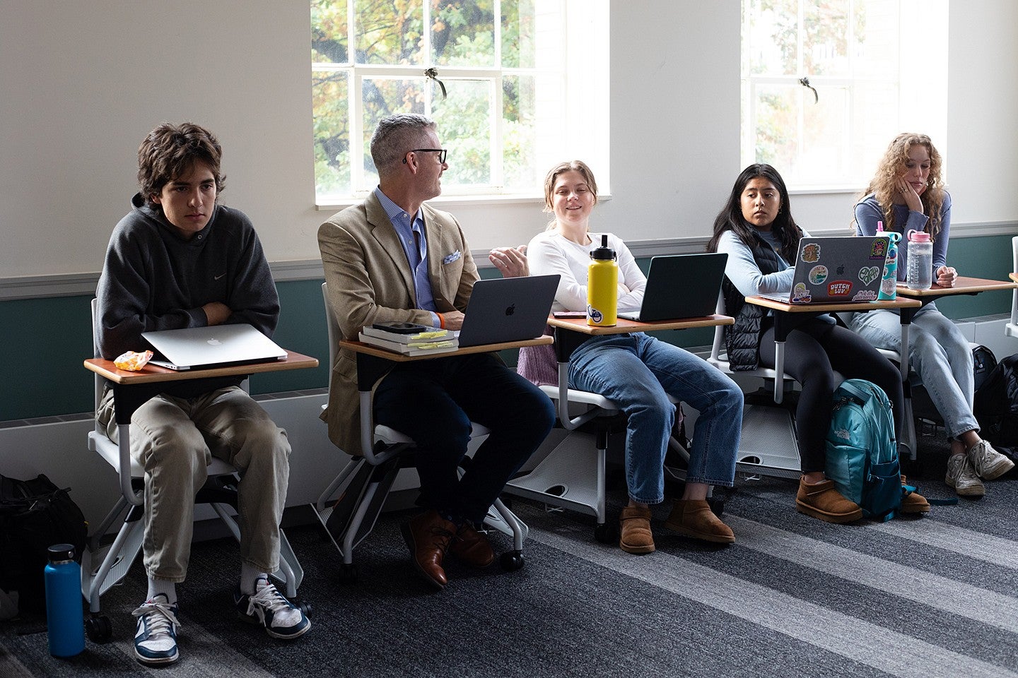 instructor tobin hansen sitting at a desk in a row of students at desks, talking to one of them