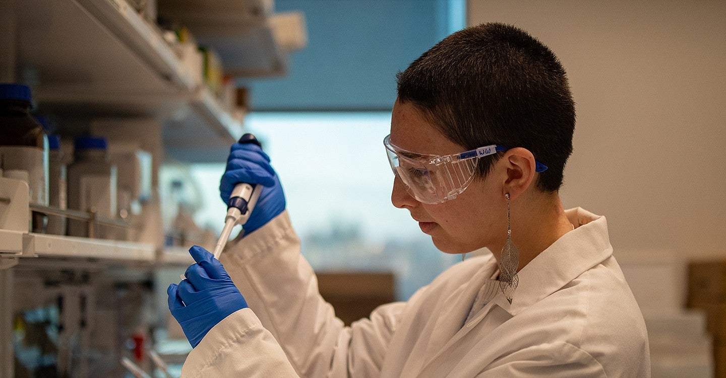 student waverly wilson in lab using pipette at bench, wearing blue gloves and white lab coat