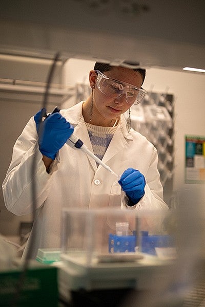 student waverly wilson in lab using pipette at bench, wearing blue gloves, white lab coat and safety glasses