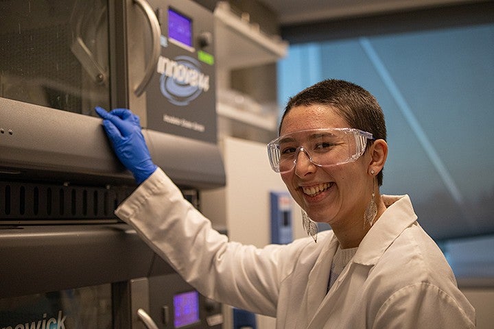 student waverly wilson smiling over shoulder at camera while working at lab bench, wearing white coat, blue gloves and safety glasses