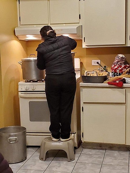 a woman standing on a stool over a large pot on the stove, making tamales