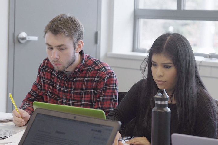 two students sitting at table, writing on paper in front of laptops