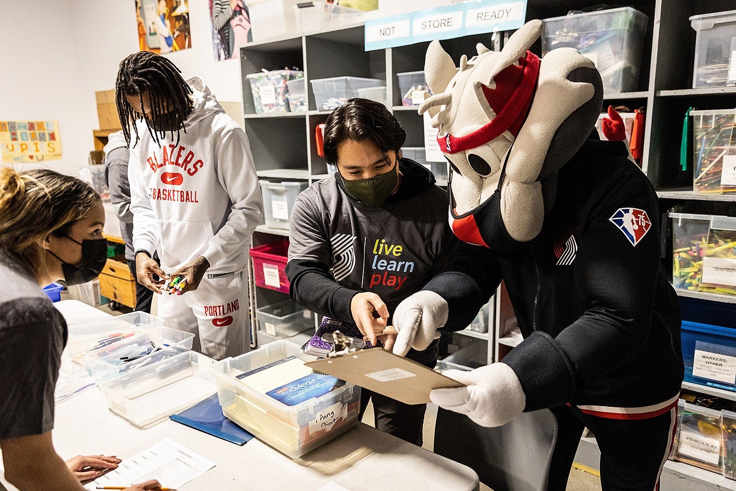 ryan horiuchi working behind the scenes with trailblazers mascot, players and other people around a table 