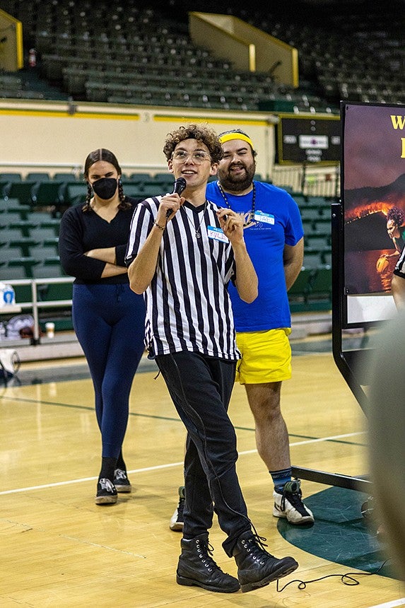 student kyle trefny wearing a referee shirt, speaking on a microphone in a basketball court