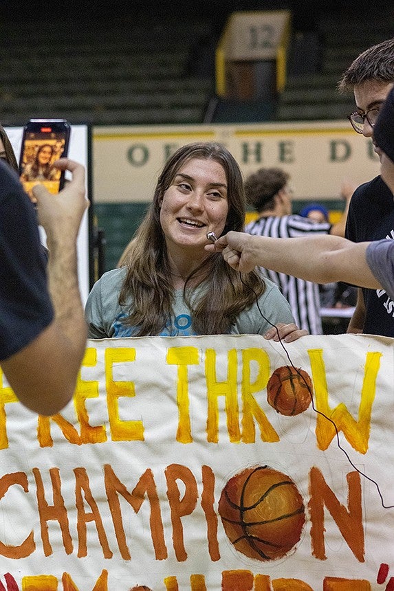 a student being interviewed and filmed on a phone while holding a hoop champion banner in an indoor basketball court