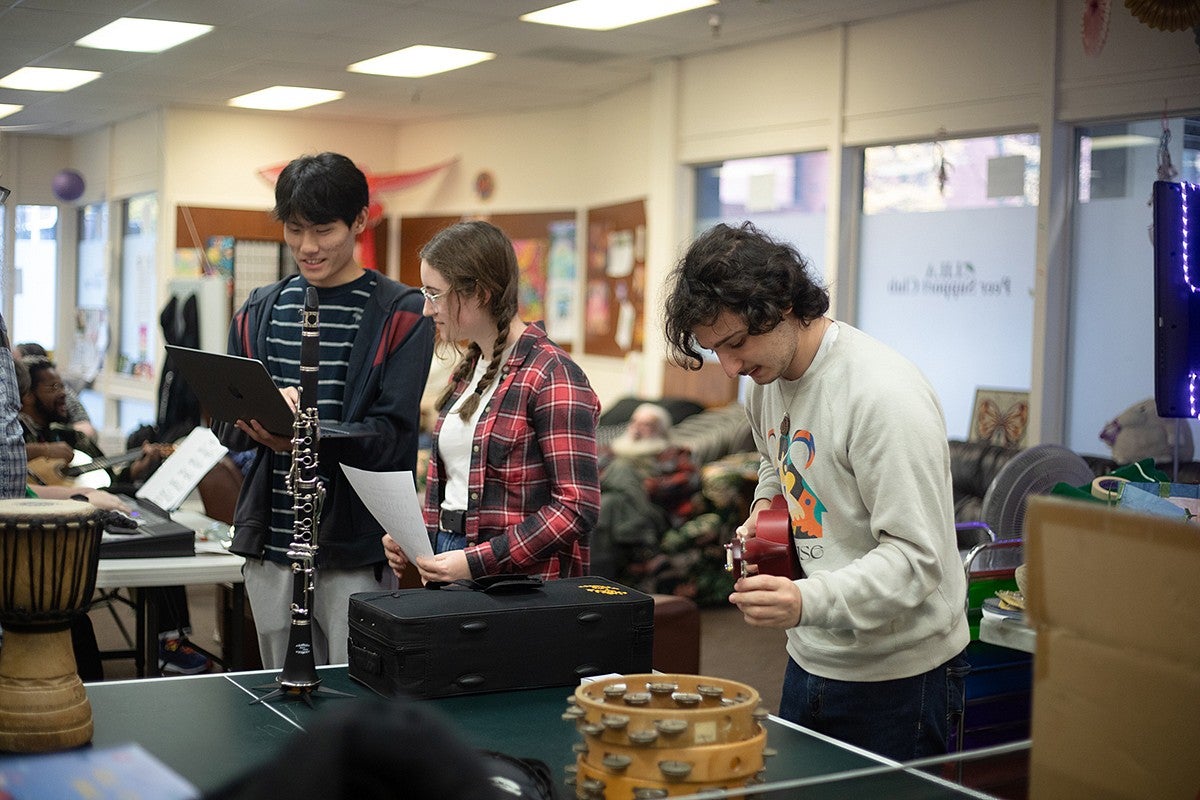 three people standing in front of table with tambourines and oboe.  One is holding a ukelele another is holding sheet music