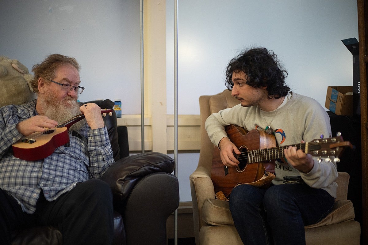 two people playing instruments, one guitar, one ukelele, sitting next to one another. 