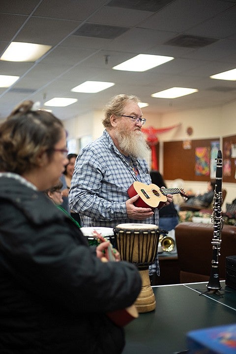 man holding ukelele in front of table with tambourines and djembe. another ukelele player in foreground. 