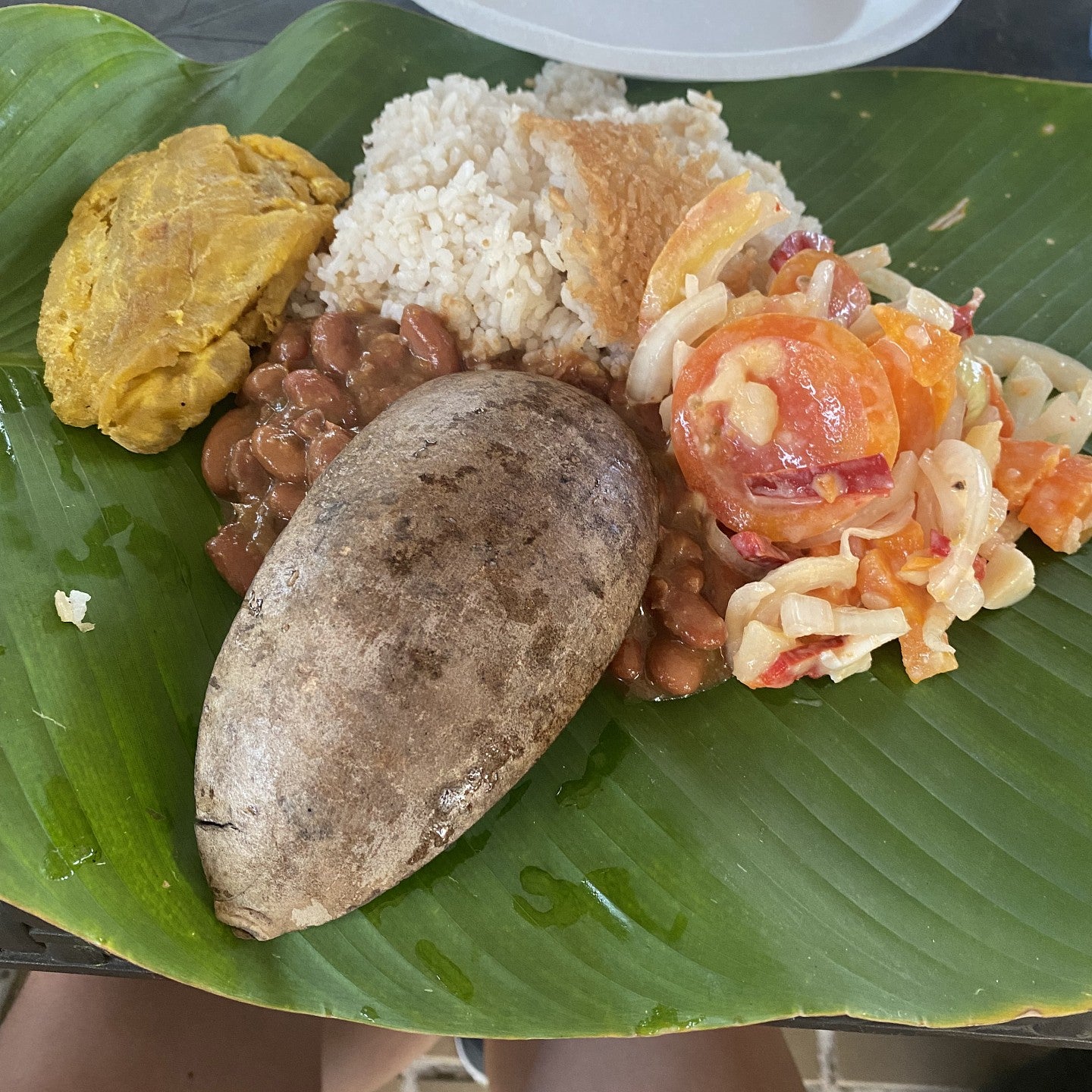 a meal of rice, beans, fried plantains, tomato salad and a baked potato nestled in a banana leaf