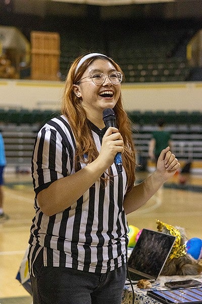 student dante' james in a referee outfit, smiling and talking into a microphone in a basketball arena