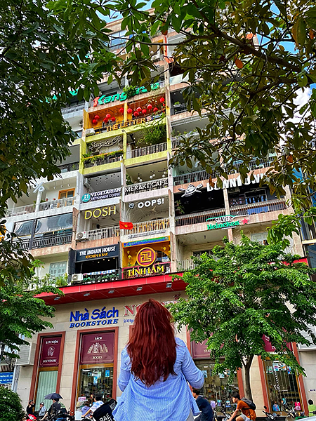 dante with back facing camera, looking up at vietnamese apartment building with colorful signage and cafe on ground floor