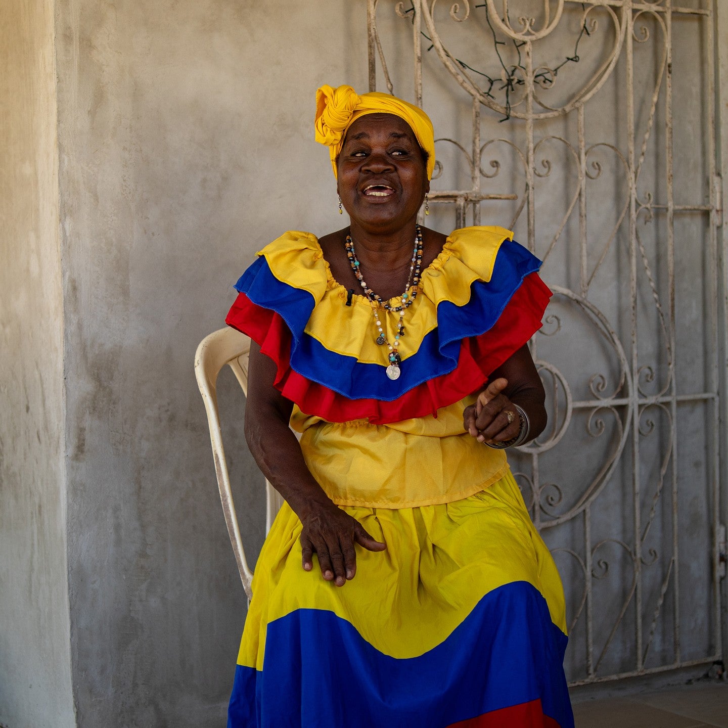 woman in brightly colored dress and headwrap seated outside stucco house, talking and gesturing