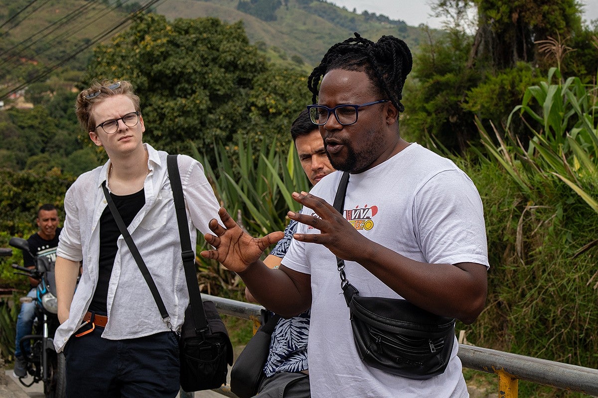 man speaking and gesturing while people next to him listen, standing along a roadside in tree-covered hills