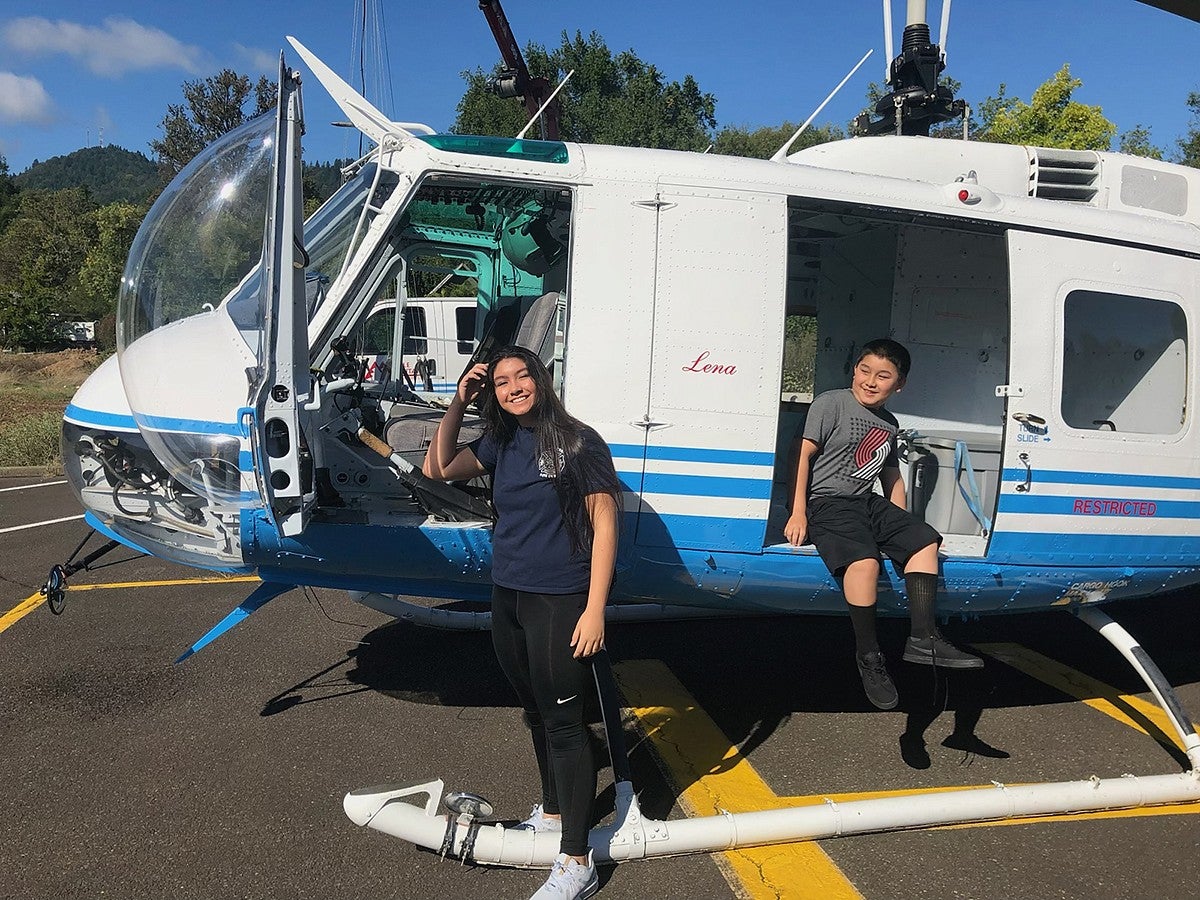 a boy and a girl posing with a helicopter parked on asphalt, with its doors open