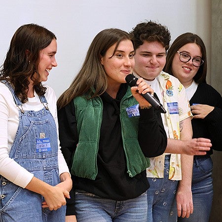 group of students standing at edge of room, one holding microphone with the others looking at her