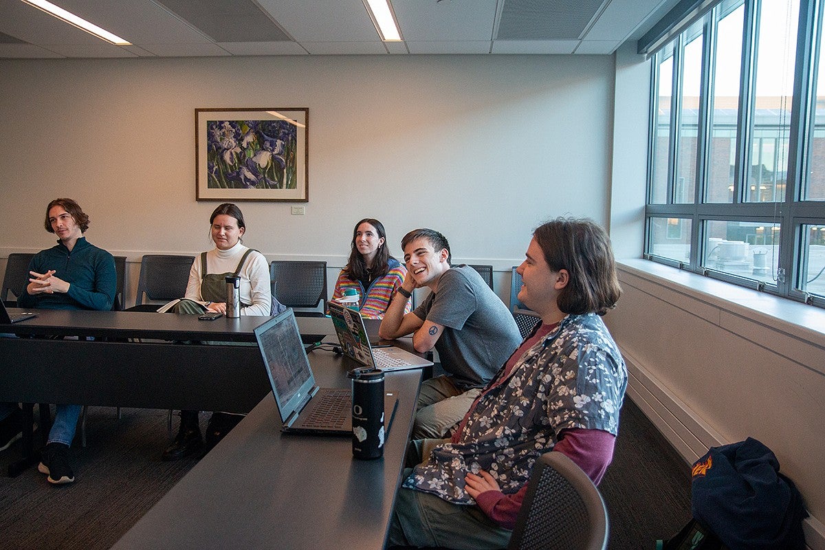 a group of students sitting at meeting tables with laptops, talking and smiling