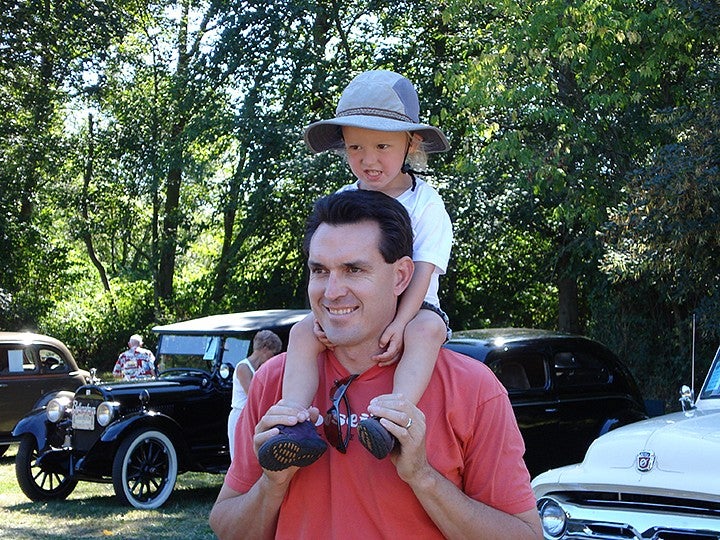a blond child on the shoulders of a man with dark hair, standing in front of antique cars parked in a grassy area with trees