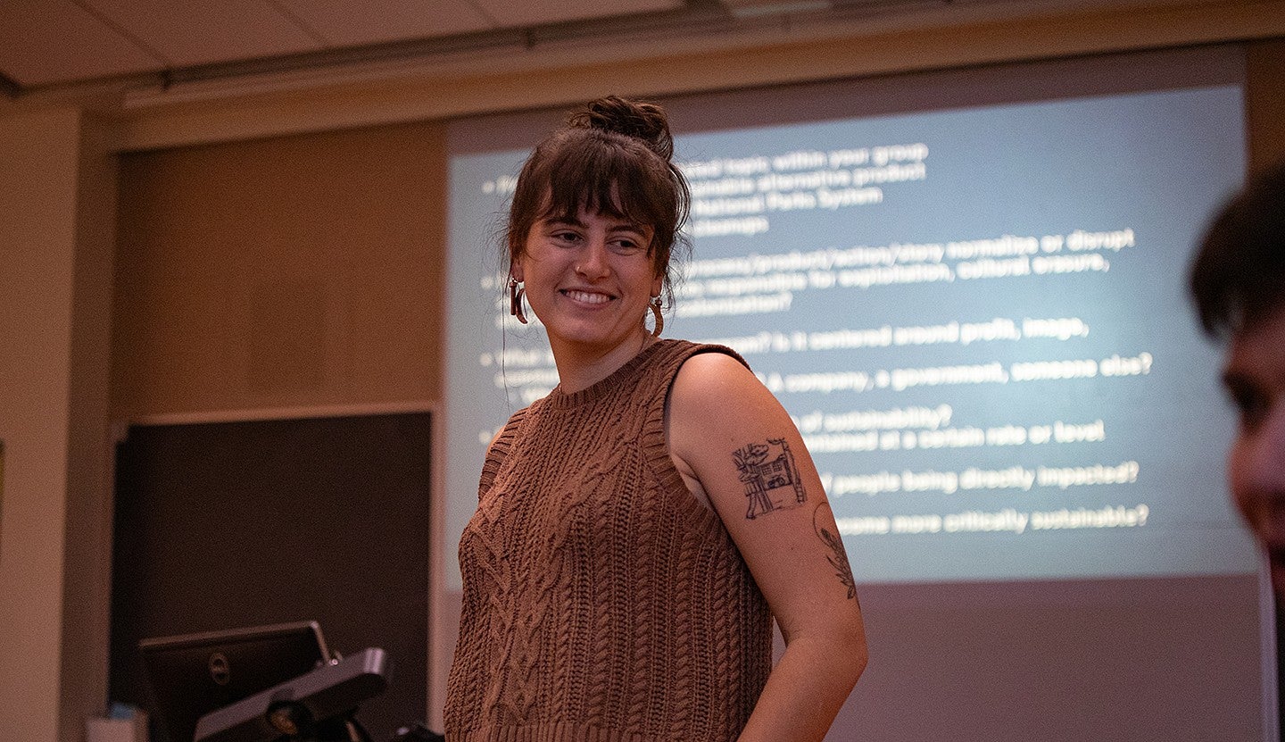 student standing at front of classroom in front of projector screen with words, smiling at another student looking on