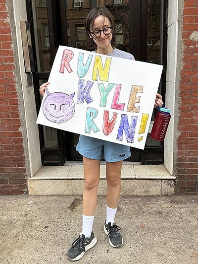 person on sidewalk holding a diy sign reading "RUN KYLE RUN" with a smiling devil emoji