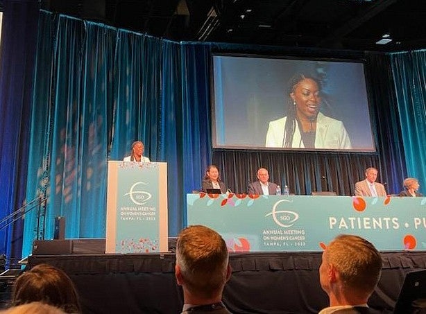 ann oluloro giving a speech at a conference, standing at podium on stage with herself projected on screen behind her