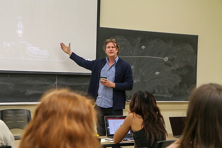 casey shoop teaching at the front of a classroom, gesturing at overhead projector screen with thermos in hand