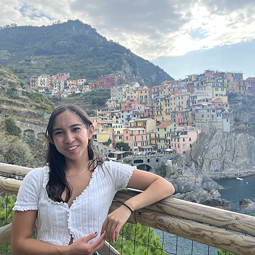 female student posing on railing with italian village on mountainous hillside overlooking ocean in background