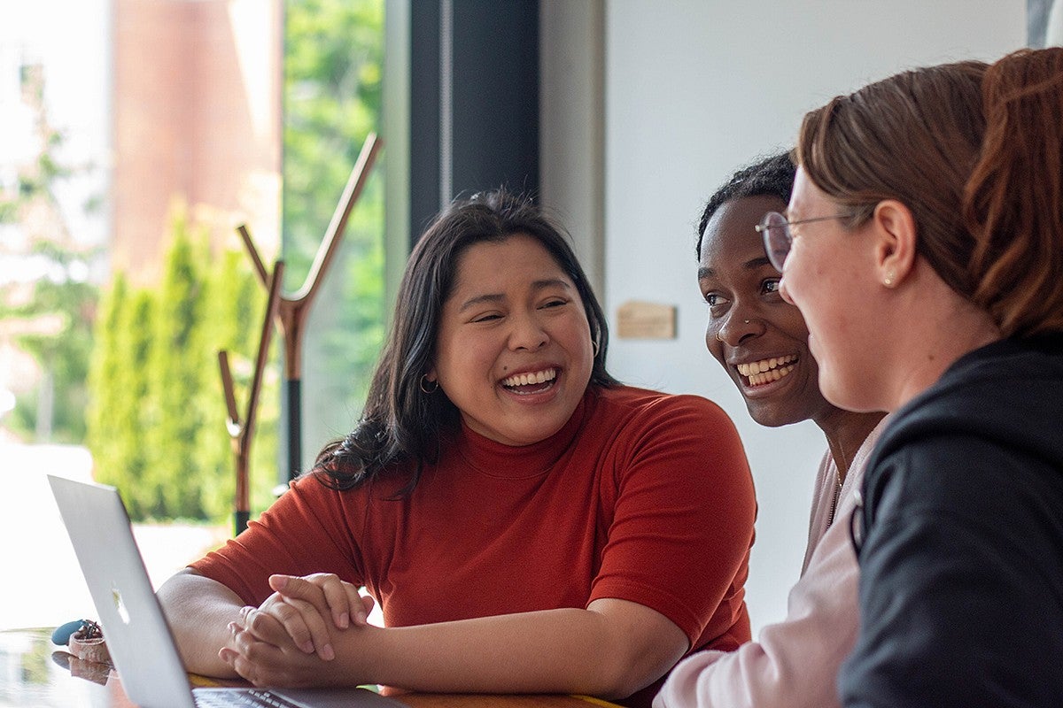 students seated in campus building at table with laptop, exchanging a laugh