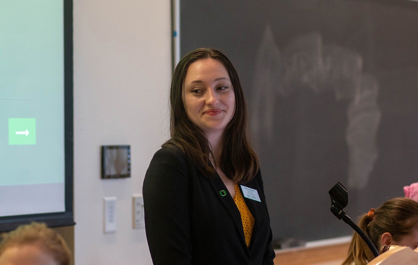 student in blazer with O pin and nametag standing in front of presentation screen, smiling at other students seated around her