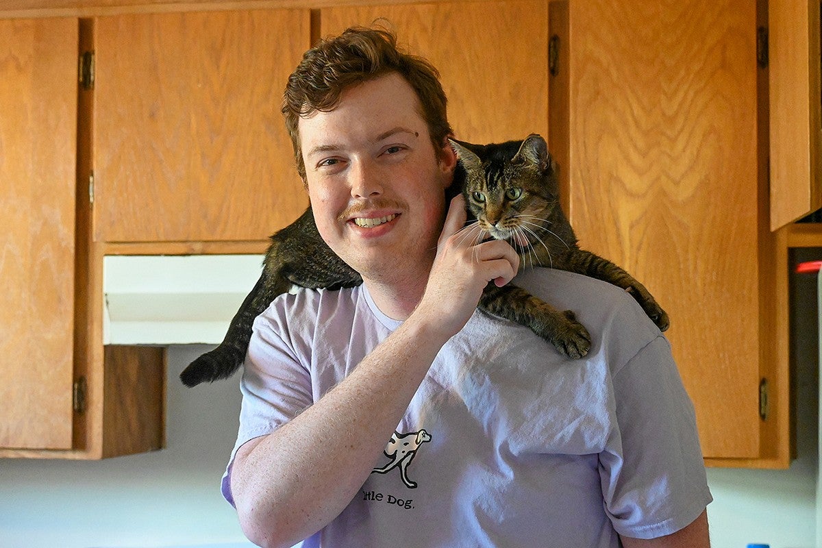 person standing in kitchen with cat on shoulders, smiling