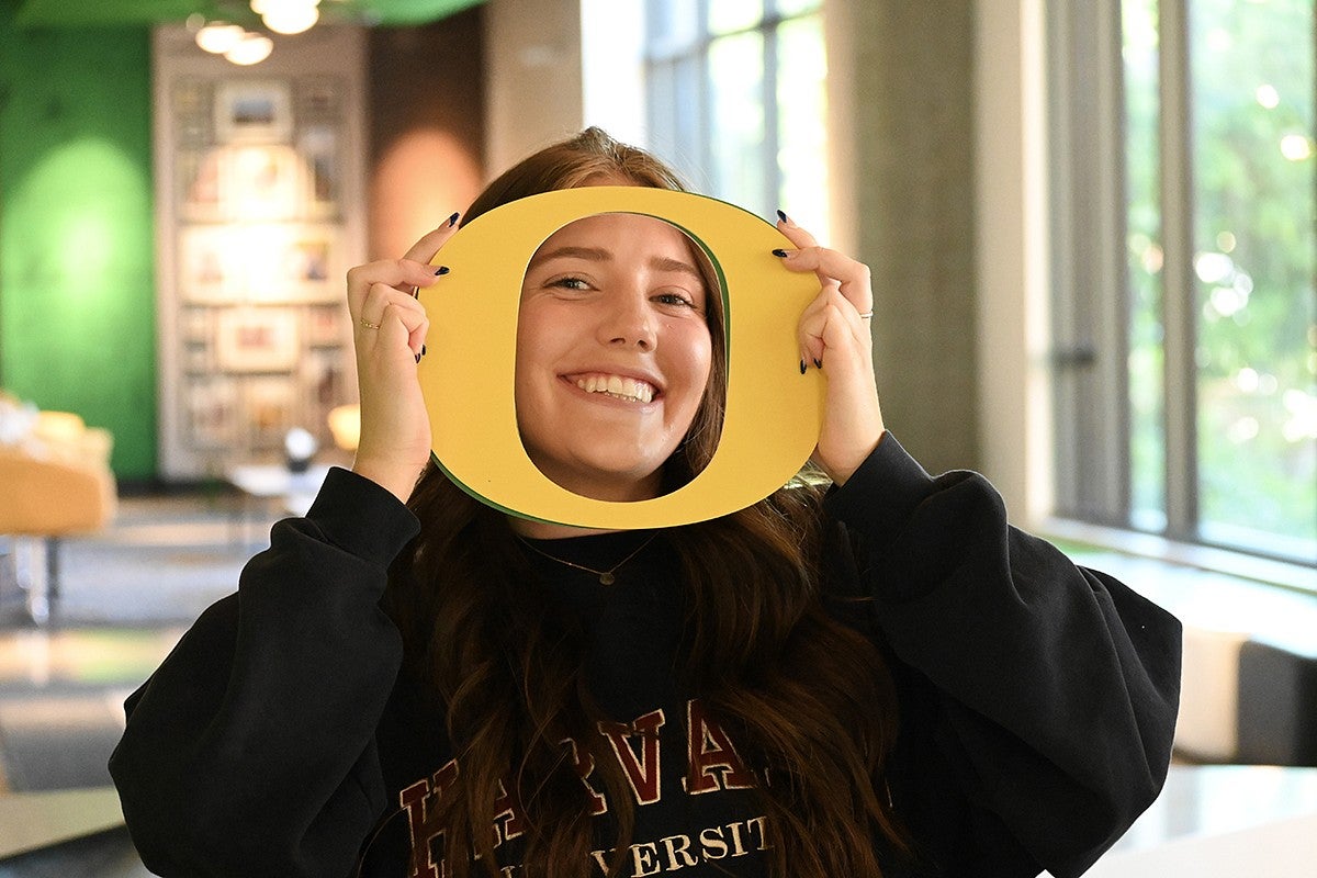 student in campus building holding yellow cutout O around their face, smiling