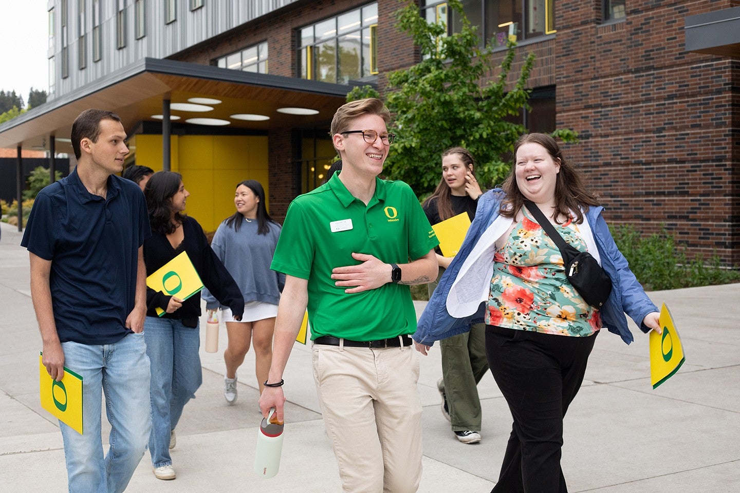 student leading campus tour, laughing with tour participants holding yellow folders and walking past a building