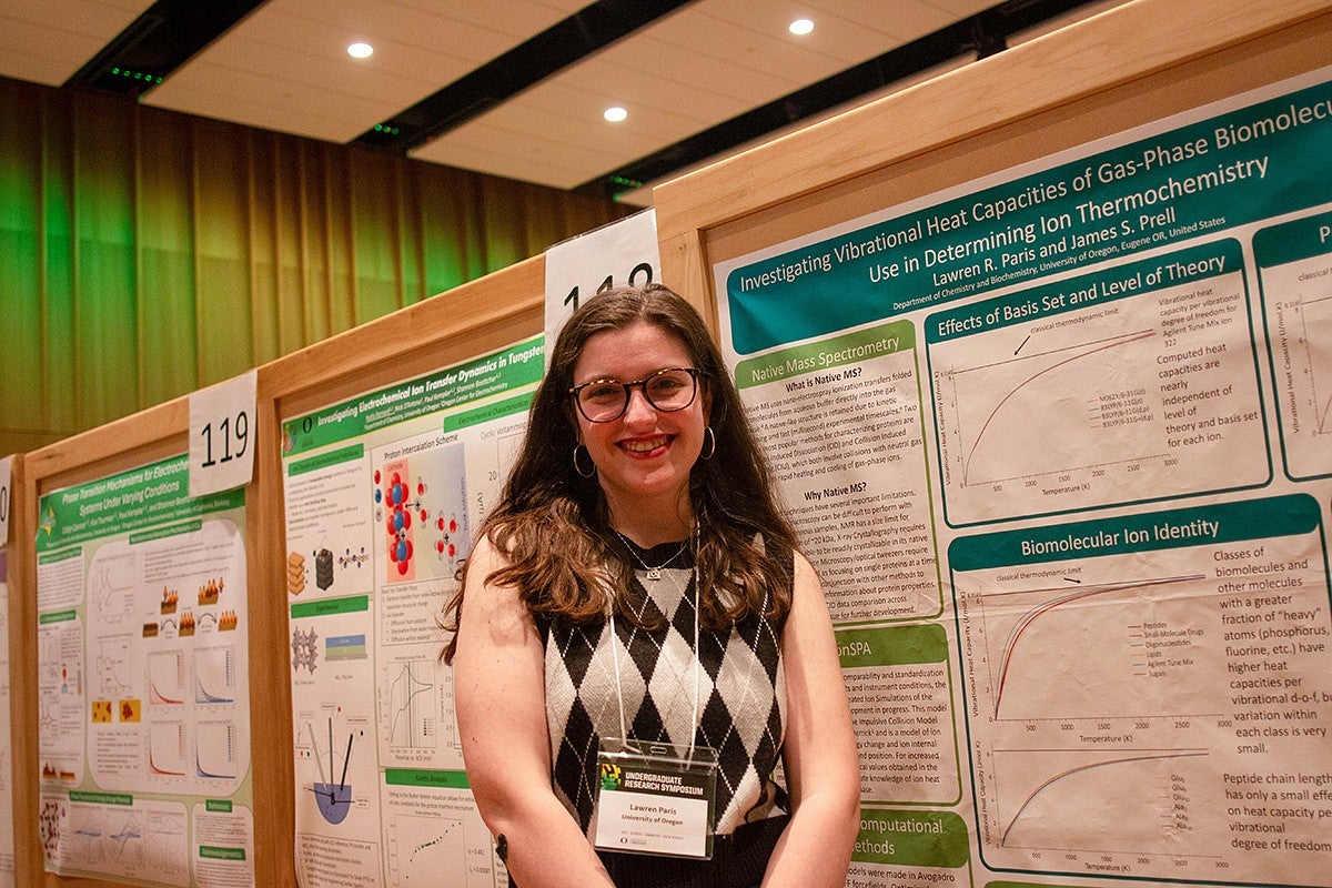 student with presenter nametag posing with chemistry poster at Undergraduate Research Symposium in campus ballroom