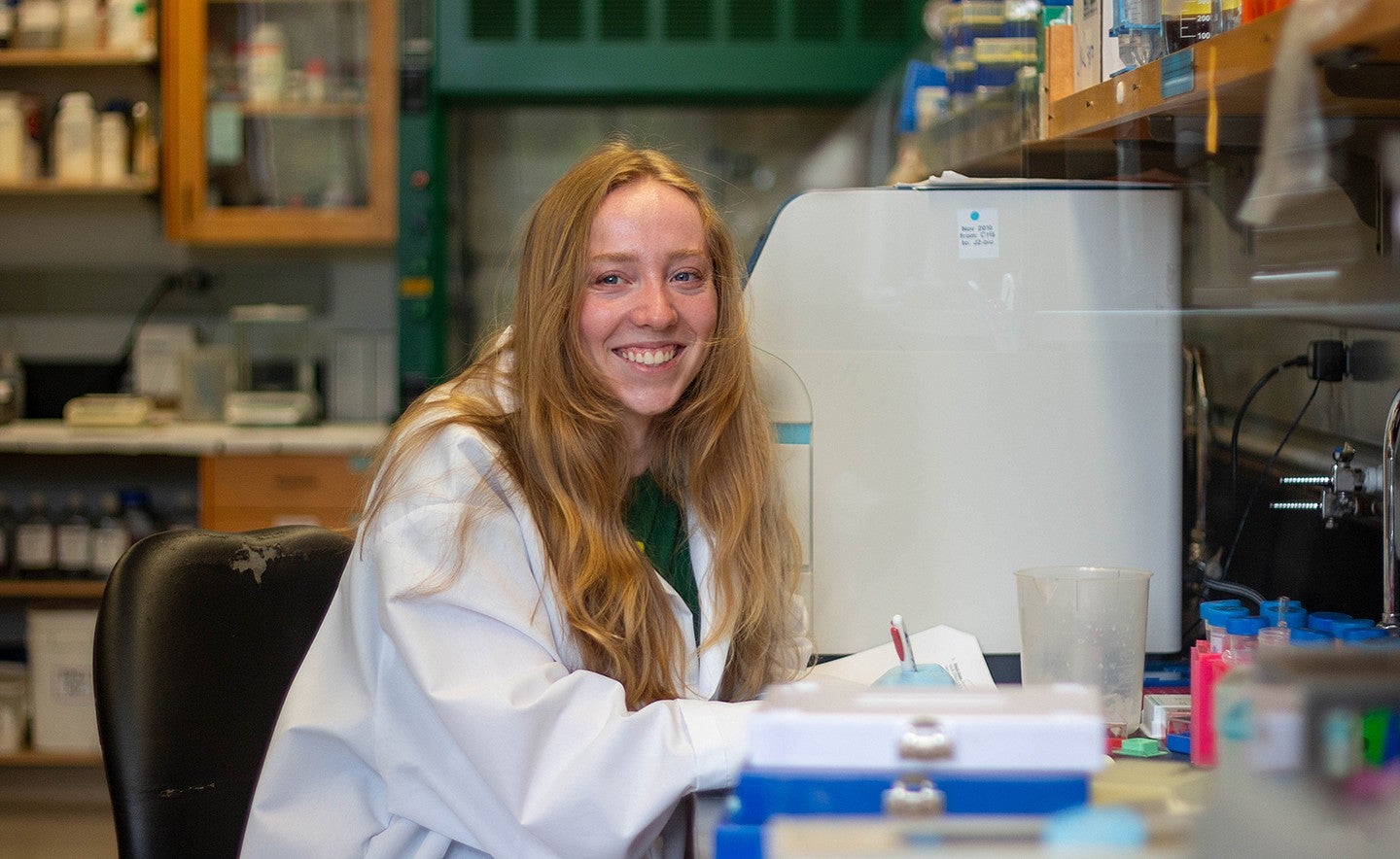 student in lab coat seated at lab bench, holding pen and smiling over her shoulder