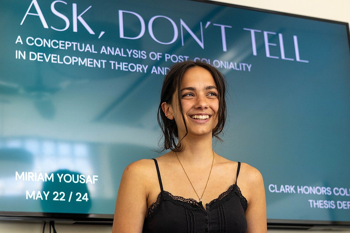 student in front of digital screen with thesis defense title slide on it reading "ask, don't tell: a conceptual analysis of post-coloniality in development theory and practice" 