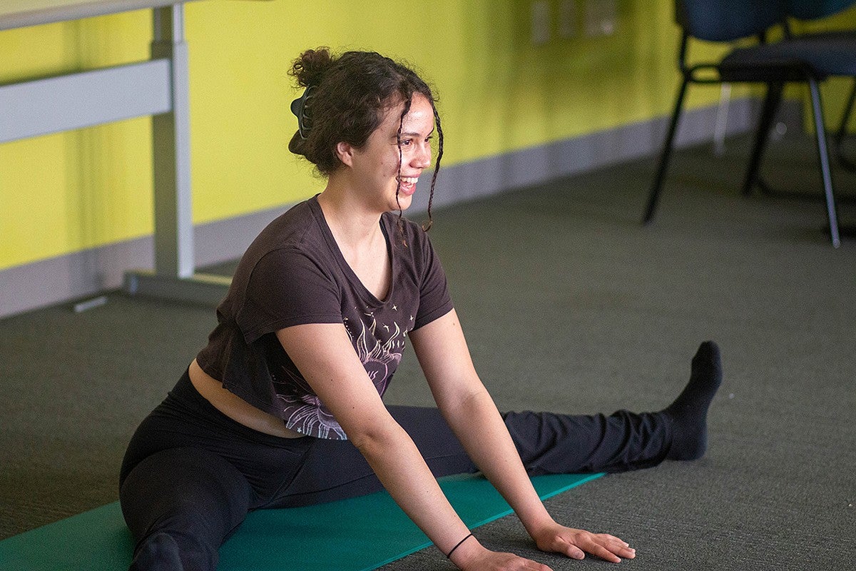 student in yoga attire sitting on mat with legs straddled, leaning forward and smiling