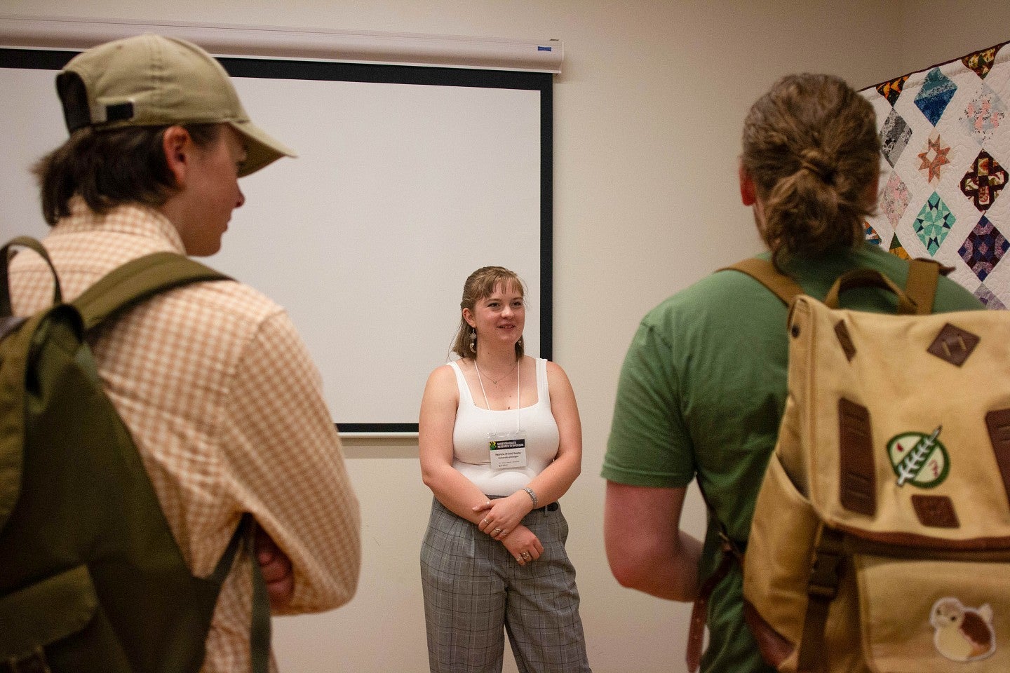 trish young wearing presenter nametag, standing in room with quilt on display, talking to two students about it